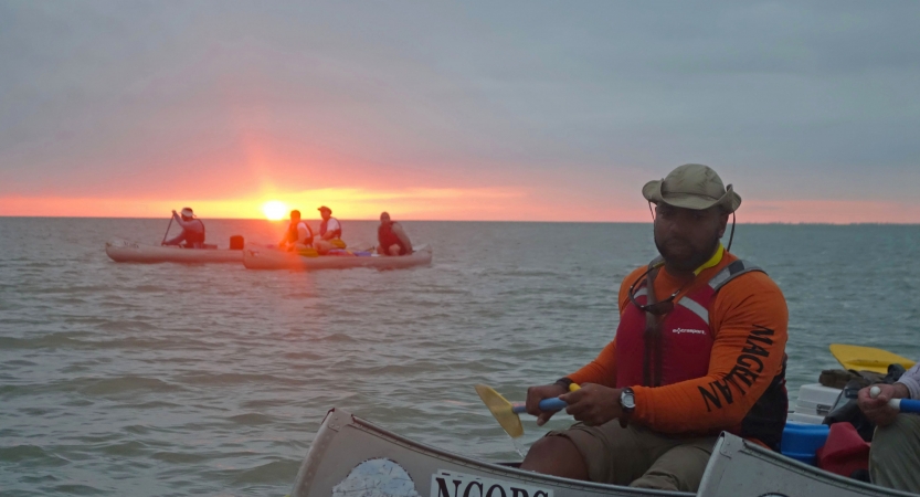a group of people paddle canoes on open water as the sun sets on the horizon